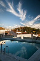 A swimming pool with a stone wall and metal railings, surrounded by buildings with red-tiled roofs and a clear sky.