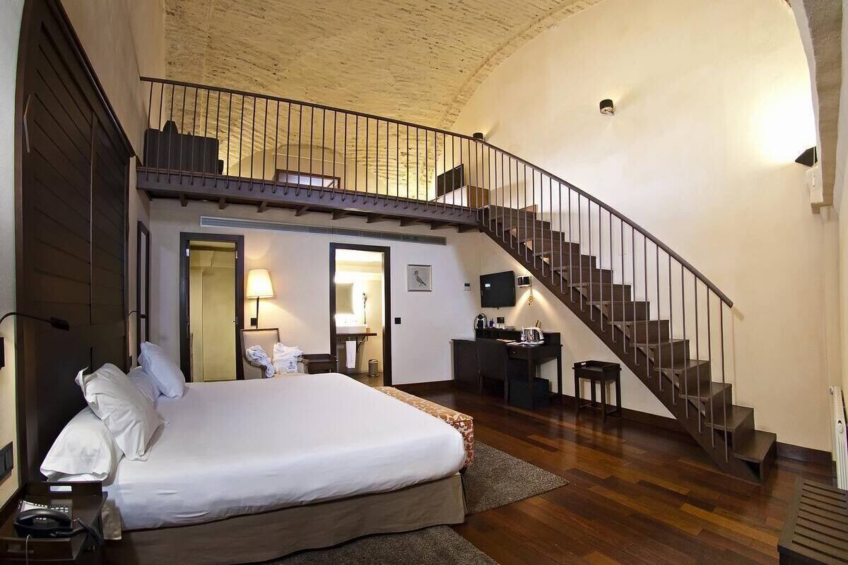 A hotel room with a large bed, a desk, a chair, a small table, a TV, and a staircase.
