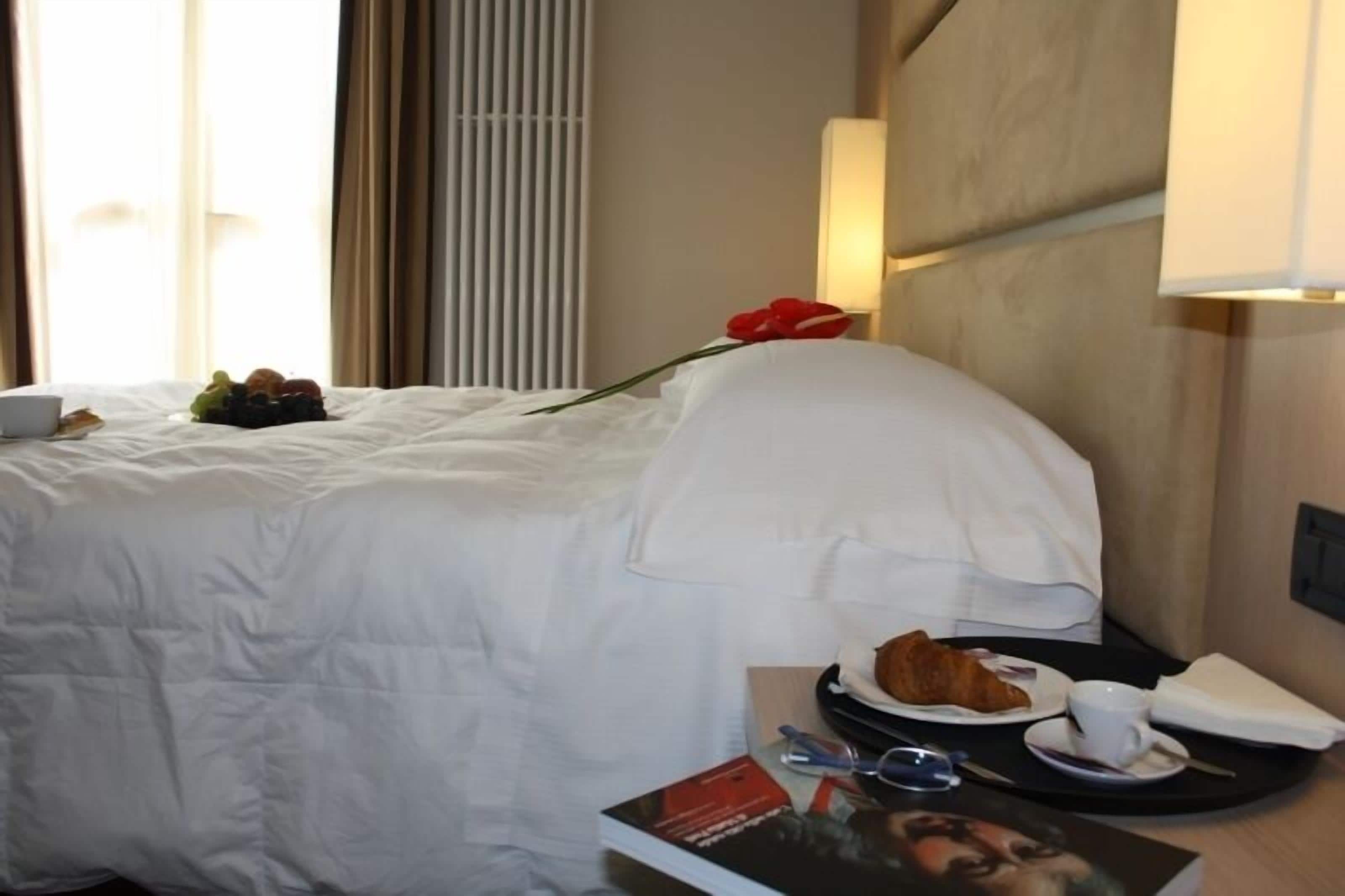 Ein Hotelzimmer mit einem ordentlich bezogenen Bett, einem Tablett mit Croissants, einer Tasse und einem Buch.