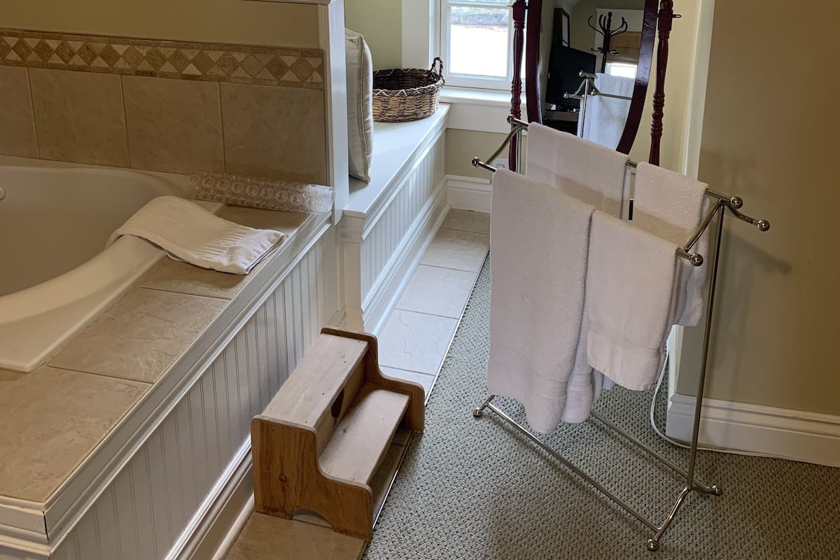 A bathroom with a bathtub, a wooden step stool, a towel rack, and a window with a view of trees.
