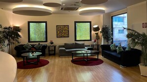 A modern living room with a sofa, two armchairs, a coffee table, and a side table. There are two windows with patterned curtains, a potted plant, and a lamp.