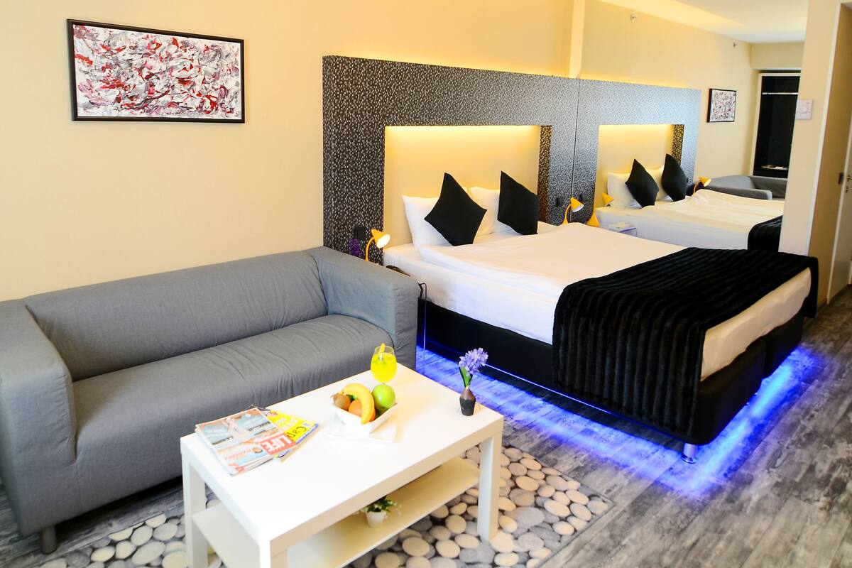 Een moderne hotelkamer met een grijze bank, een witte salontafel met tijdschriften en fruit, een bed met zwart beddengoed en blauwe verlichting onder het bed, en een groot abstract schilderij aan de muur.