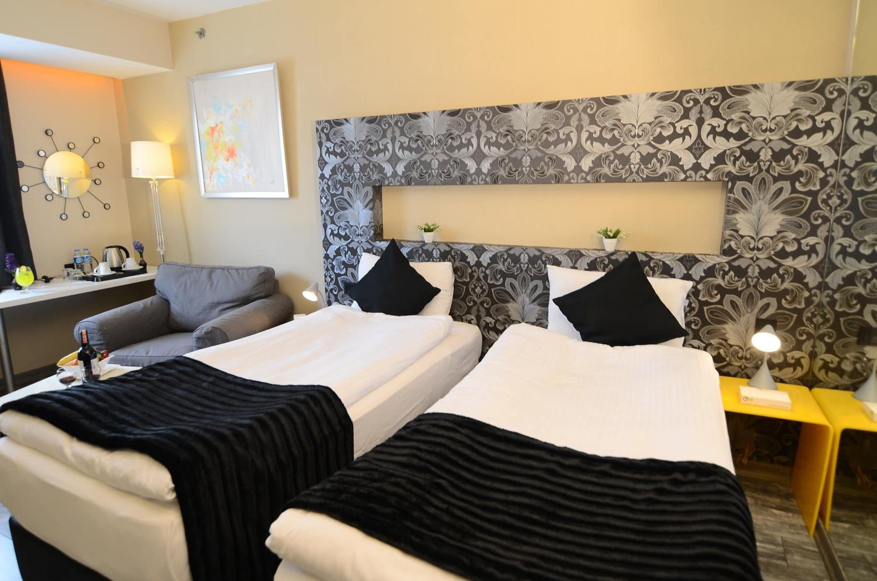 A hotel room with two beds, a sofa, a desk with a lamp, and a decorative wall.