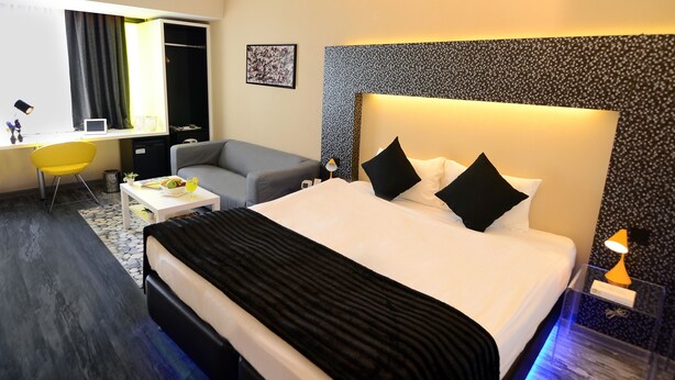A modern hotel room with a large bed, a grey sofa, a small table with a bowl of fruit, and a desk with a chair.