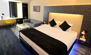 Una habitación de hotel moderna con una cama grande, un sofá gris, una mesita con un frutero y un escritorio con una silla.