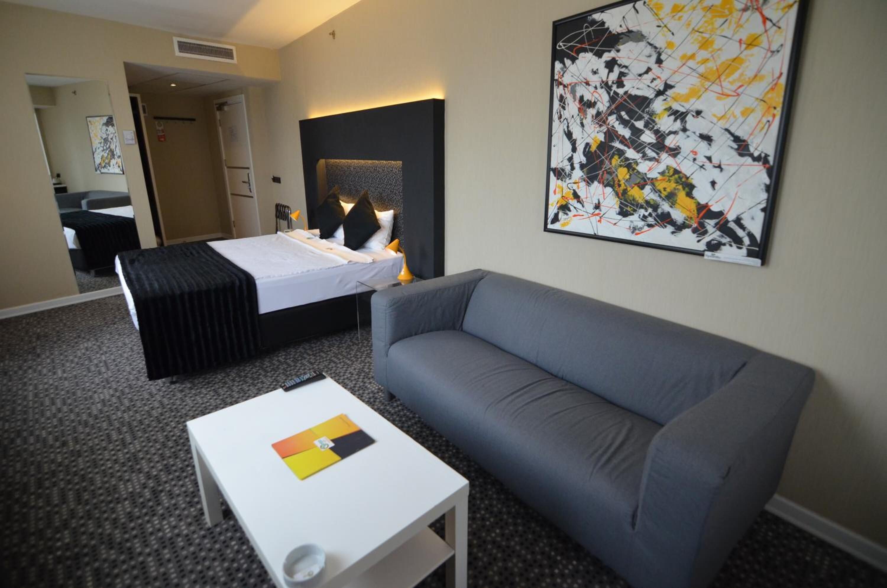 A hotel room with a bed, a gray sofa, a white coffee table, and a large abstract painting on the wall.