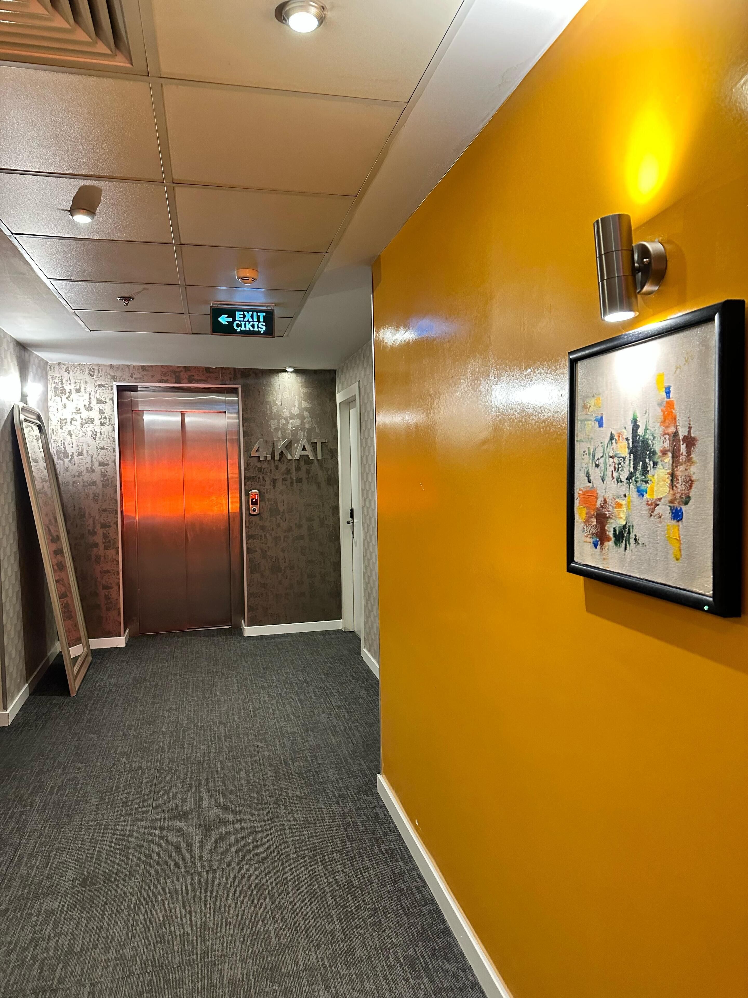 An elevator with a metallic finish, a wall-mounted exit sign, and a framed abstract painting.