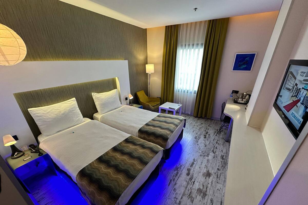 A hotel room with two beds, a desk, a chair, a lamp, and a TV displaying a map.