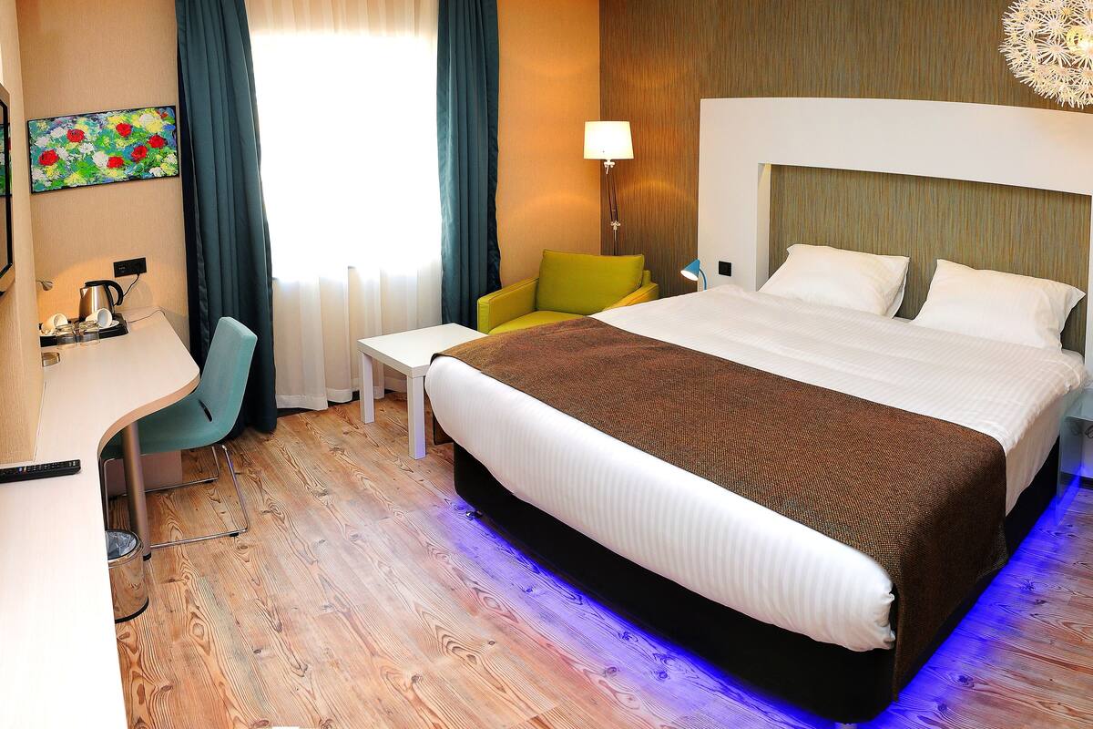 A hotel room with a large bed, a desk, a chair, a lamp, and a painting on the wall.