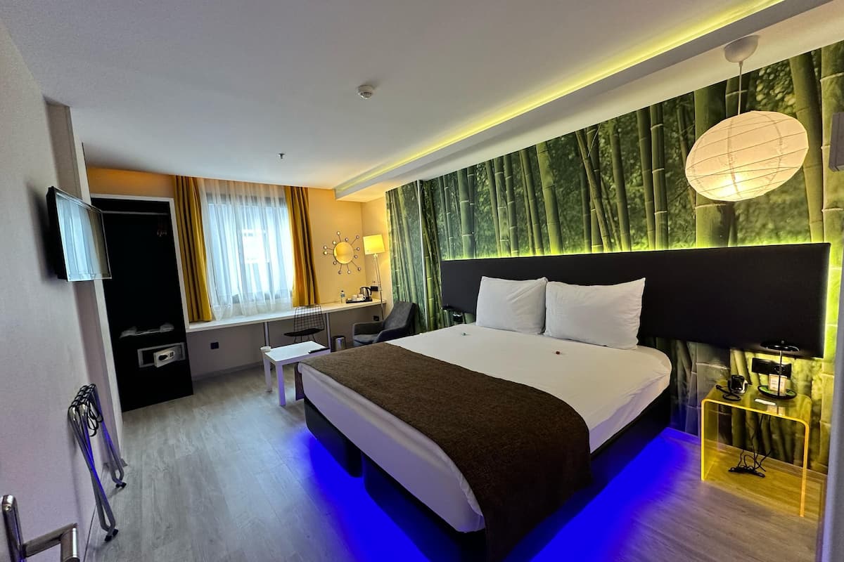 Une chambre d’hôtel moderne dotée d’un grand lit, d’un bureau avec une chaise, d’une télévision et d’un mur orné d’une fresque représentant une forêt de bambous.