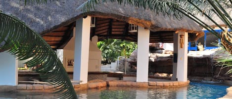 Un área de piscina con una estructura de techo de paja, hojas de palma y una piscina azul cristalina.