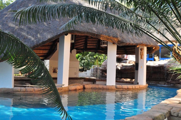 A pool area with a thatched-roof structure, palm fronds, and a clear blue pool.