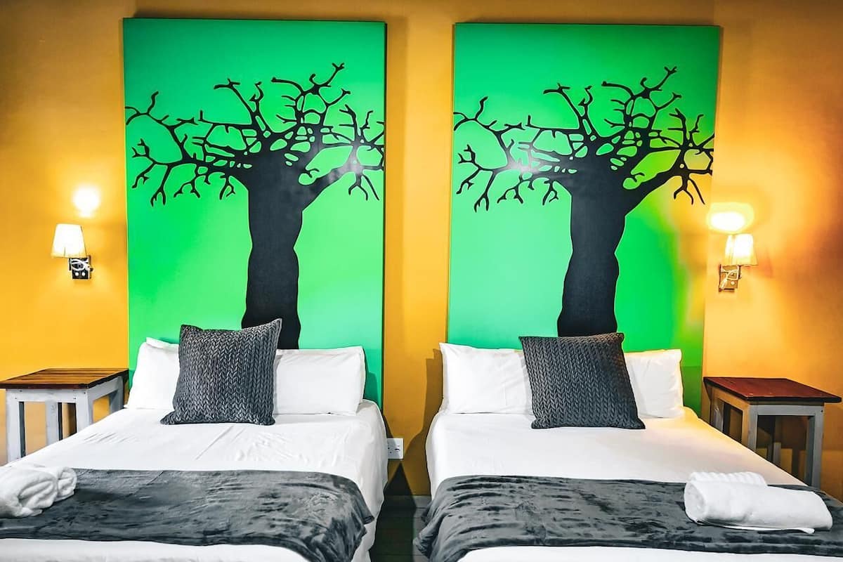 Habitación de hotel con dos camas, cada una con un cojín estampado en blanco y negro, frente a una pared verde con un mural de un árbol negro.