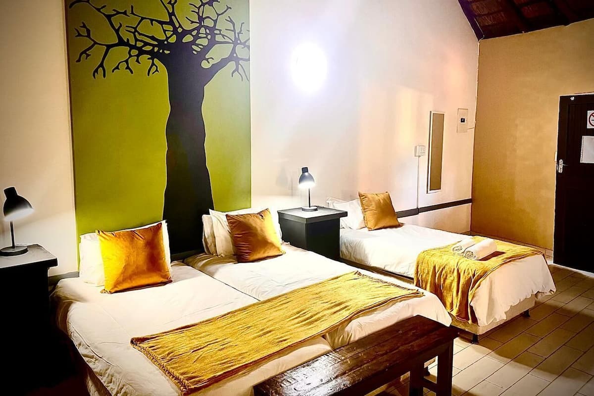 Habitación de hotel con dos camas, una mesita de noche con lámpara, un cuadro de un árbol en la pared y una puerta con un cartel.