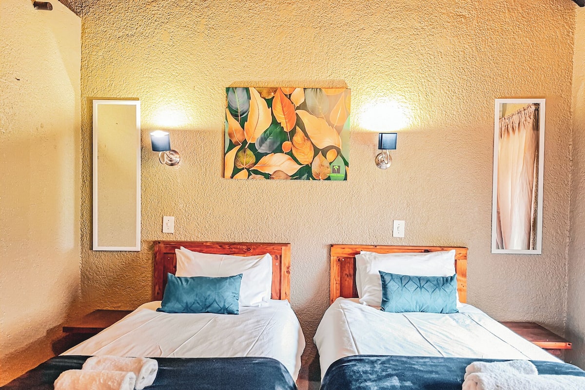Habitación de hotel con dos camas, cada una con cabecero de madera, ropa de cama blanca y almohadas azules. Hay luces fijadas a la pared y un cuadro sobre las camas.