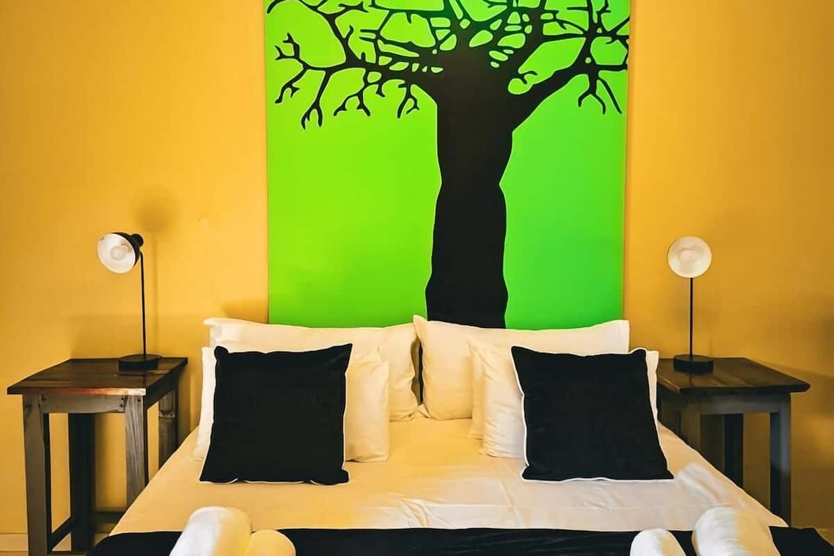 Habitación de hotel con una cama, dos almohadas negras, una colcha blanca, una pared verde y un cuadro de un árbol en blanco y negro.