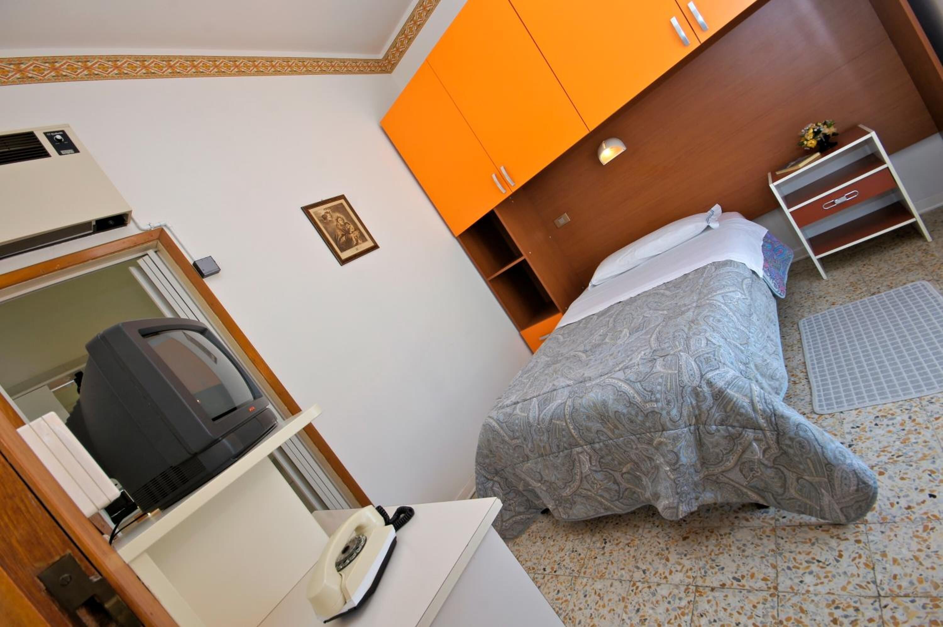 A hotel room with a bed, a desk with a telephone, an air conditioning unit, and a wardrobe.