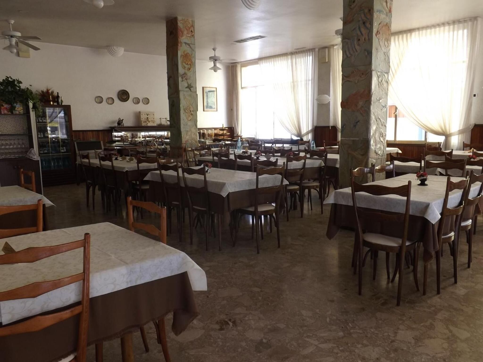 A spacious dining area with multiple tables and chairs, a bar area, and large windows with curtains.