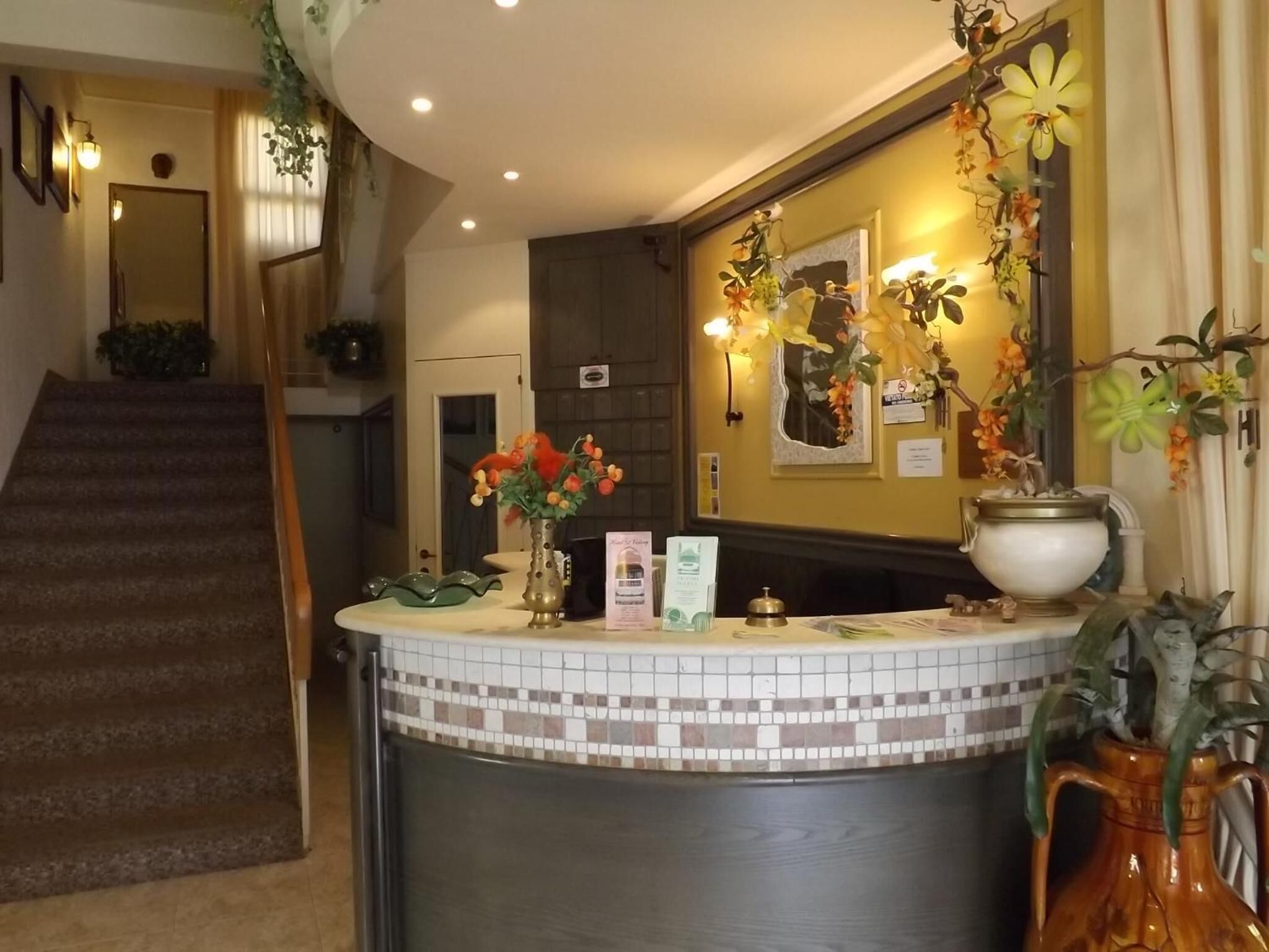 Un área de recepción con un mostrador de baldosas, un jarrón con flores, una planta en maceta y una escalera que sube a otro piso.