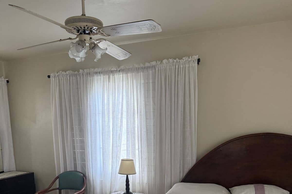 Habitación de hotel con cama, una silla, mesita de noche con lámpara, ventilador de techo y ventana con cortinas.