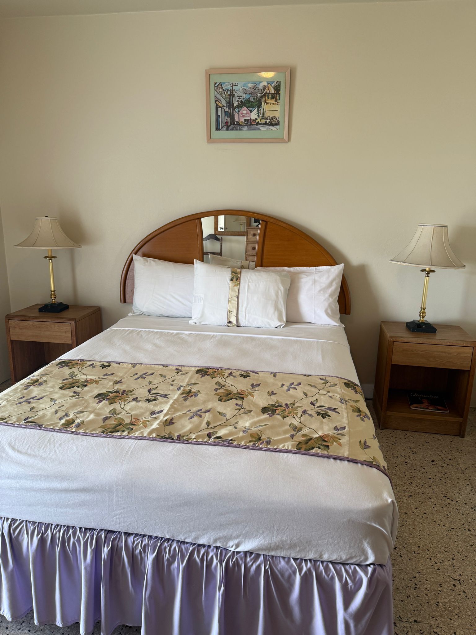 Una cama bien hecha con colcha floral y cabecera de madera. Hay dos mesitas de noche con lámparas y un cuadro enmarcado en la pared sobre la cama.