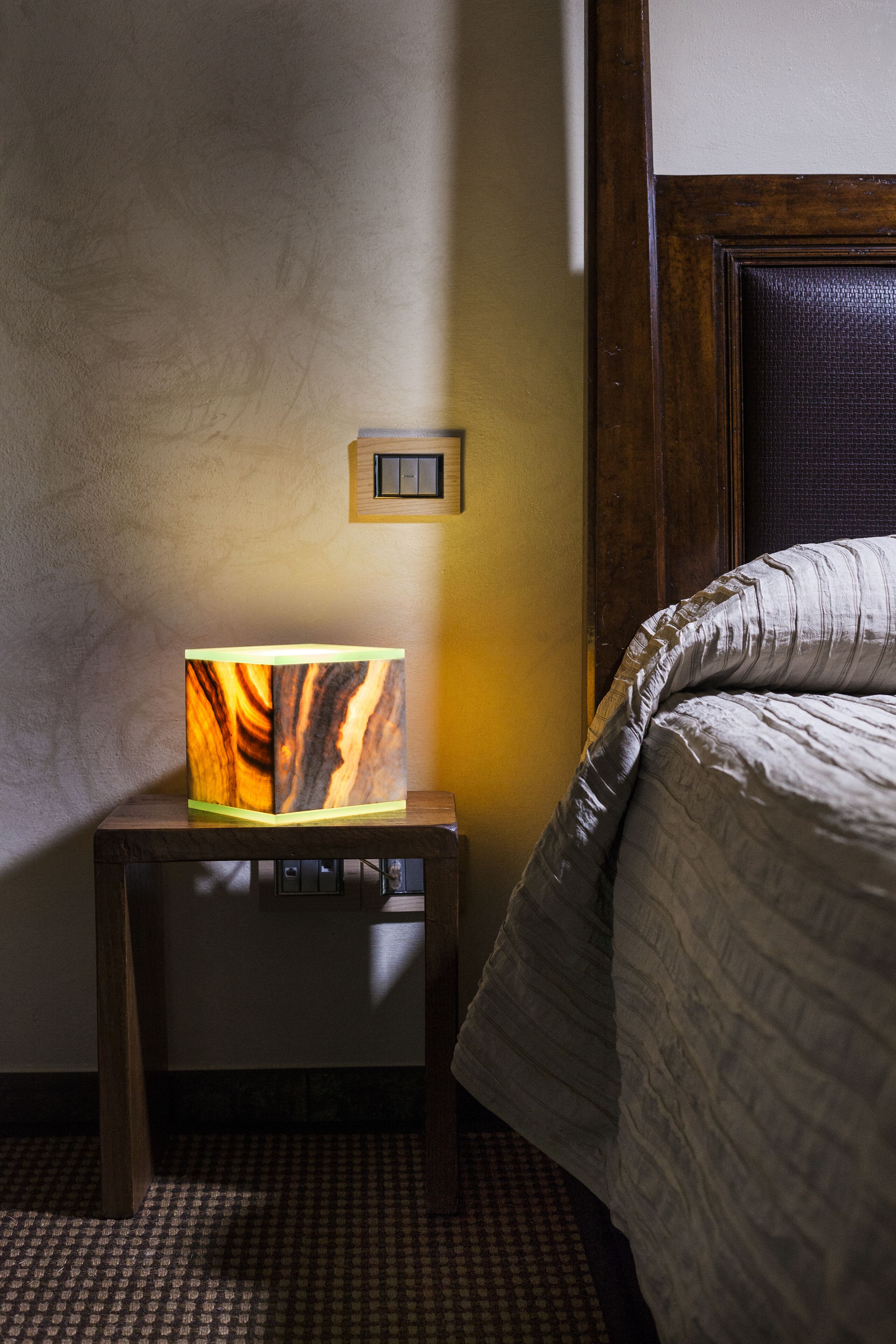 A hotel room with a bed, a nightstand with a lamp, a wall switch, and a textured wall.