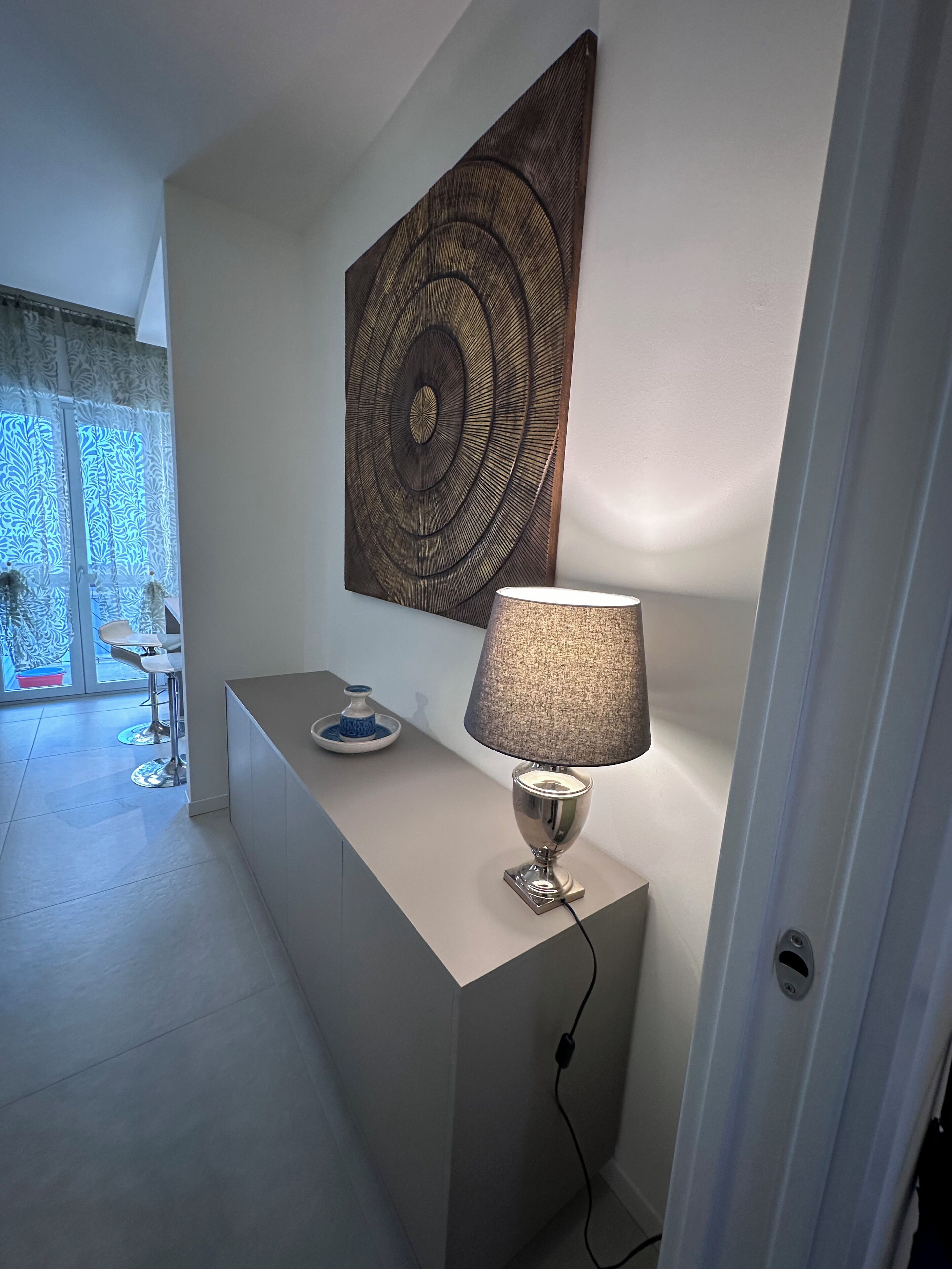 A modern interior with a minimalist design, featuring a desk with a lamp, a decorative wall piece, and a glimpse of a dining area with a patterned curtain.