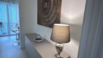 A modern interior with a minimalist design, featuring a desk with a lamp, a decorative wall piece, and a glimpse of a dining area with a patterned curtain.