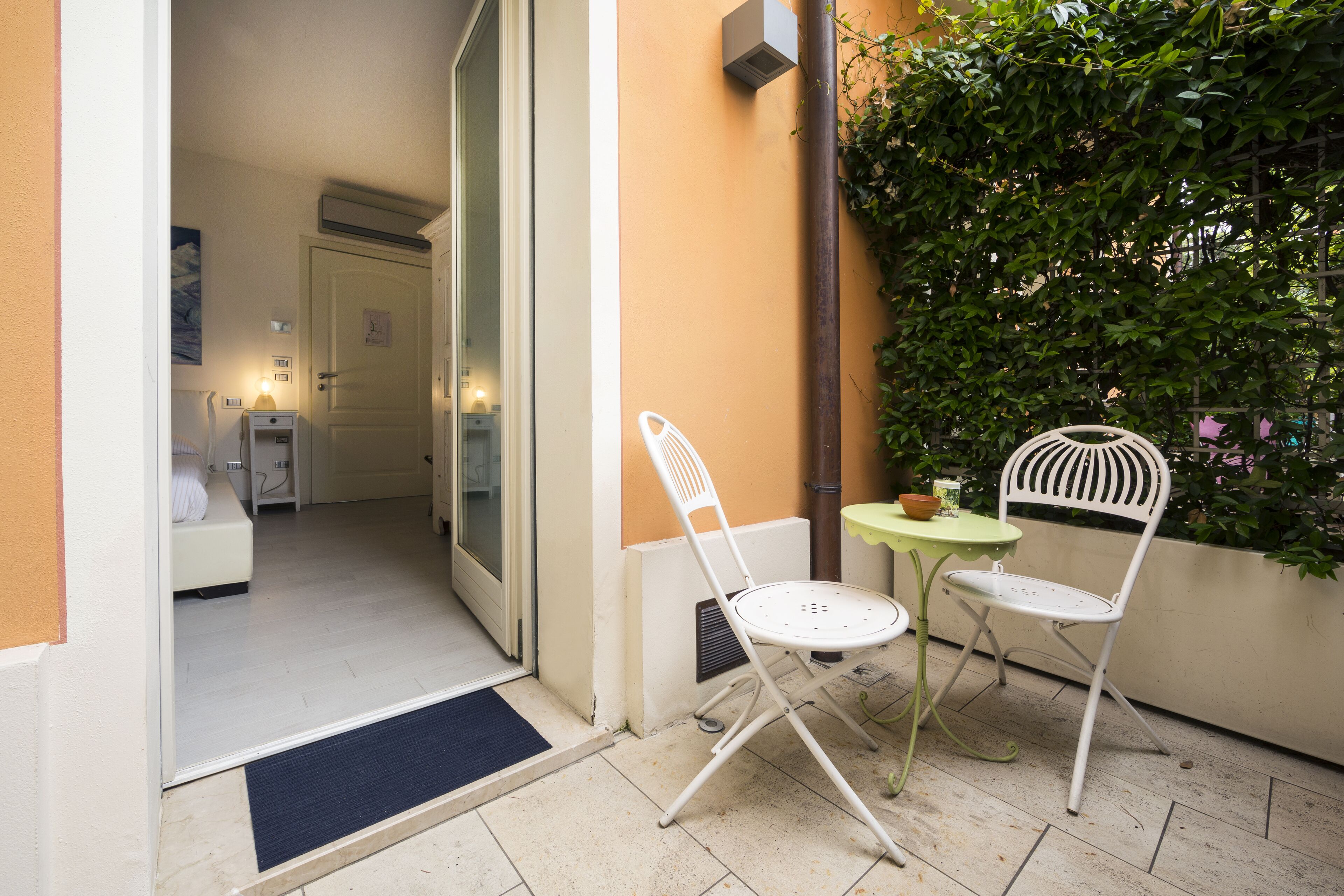 Un pequeño espacio exterior con dos sillas blancas y una mesa verde, una puerta que da a una habitación y una planta en maceta.