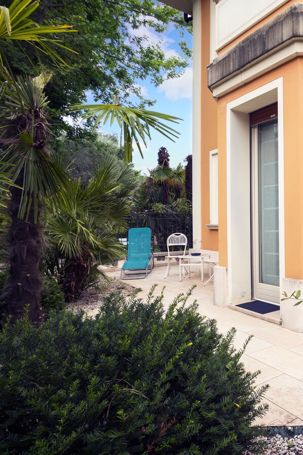 A patio with a blue chair and a white chair, surrounded by greenery and a building with a yellow facade. - La Villa Desenzano (Desenzano del Garda)