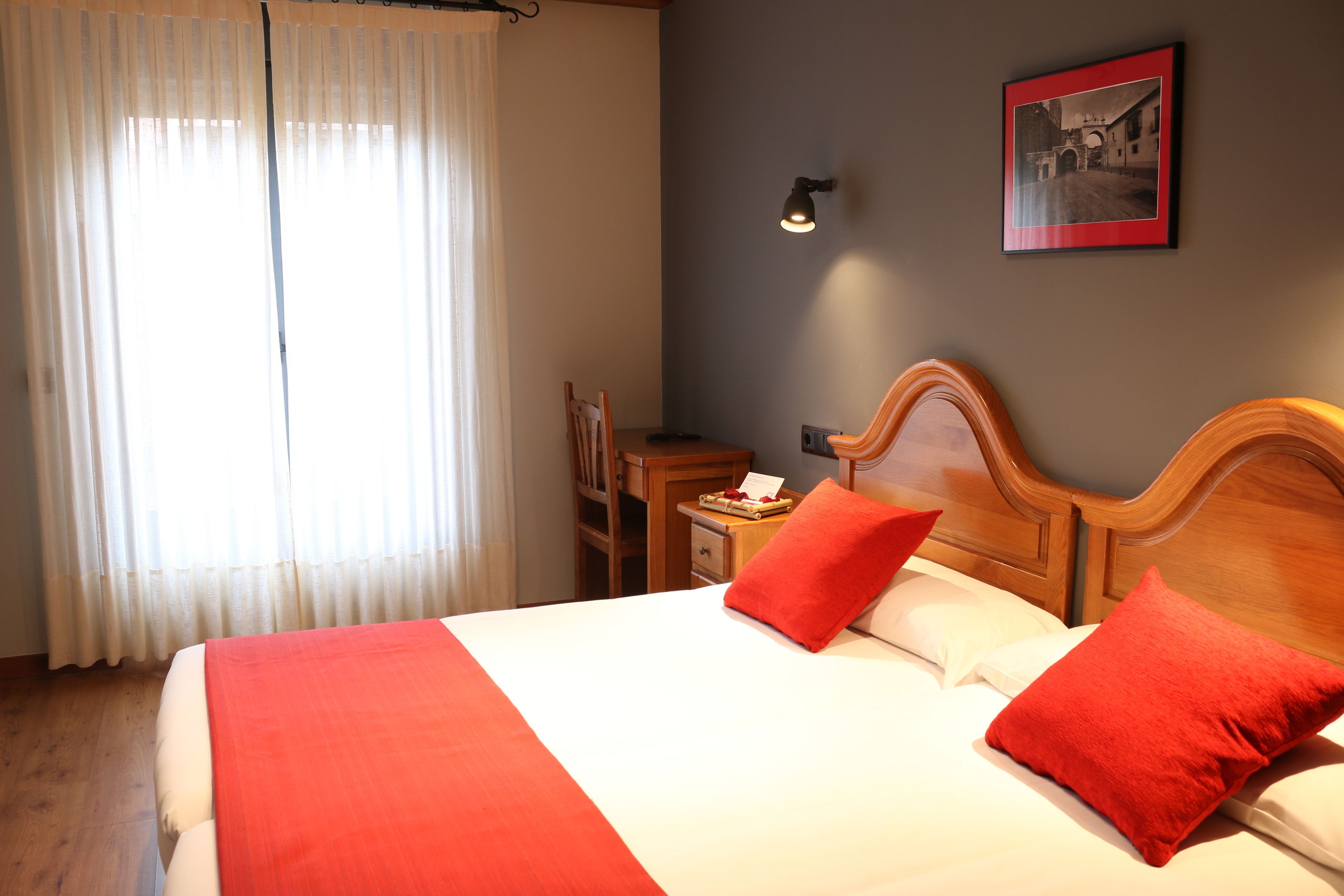 Hotelkamer met een groot bed, twee rode kussens, een houten hoofdbord, een nachtkastje met een lamp, een ingelijst schilderij aan de muur, en een raam met dunne gordijnen.