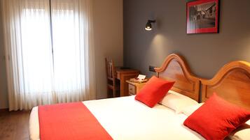 Hotelkamer met een groot bed, twee rode kussens, een houten hoofdbord, een nachtkastje met een lamp, een ingelijst schilderij aan de muur, en een raam met dunne gordijnen.
