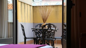 Ein Zimmer mit einem Bett, einem Tisch und Stühlen. Ein Fenster mit Vorhängen ist zu sehen, und eine Tür ist ebenfalls erkennbar.