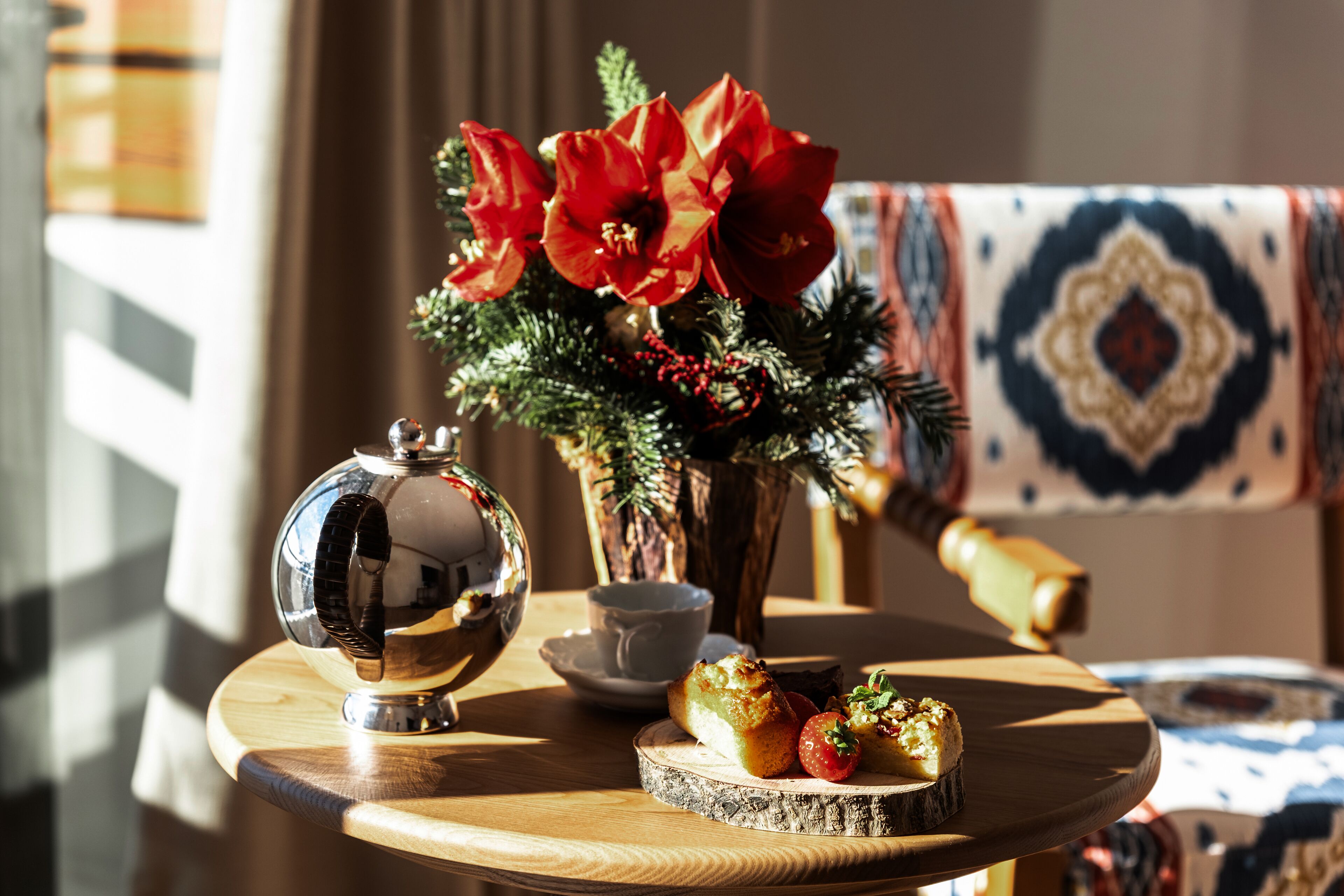 A round wooden table with a teapot, a cup, a plate with food, and a vase with red flowers.