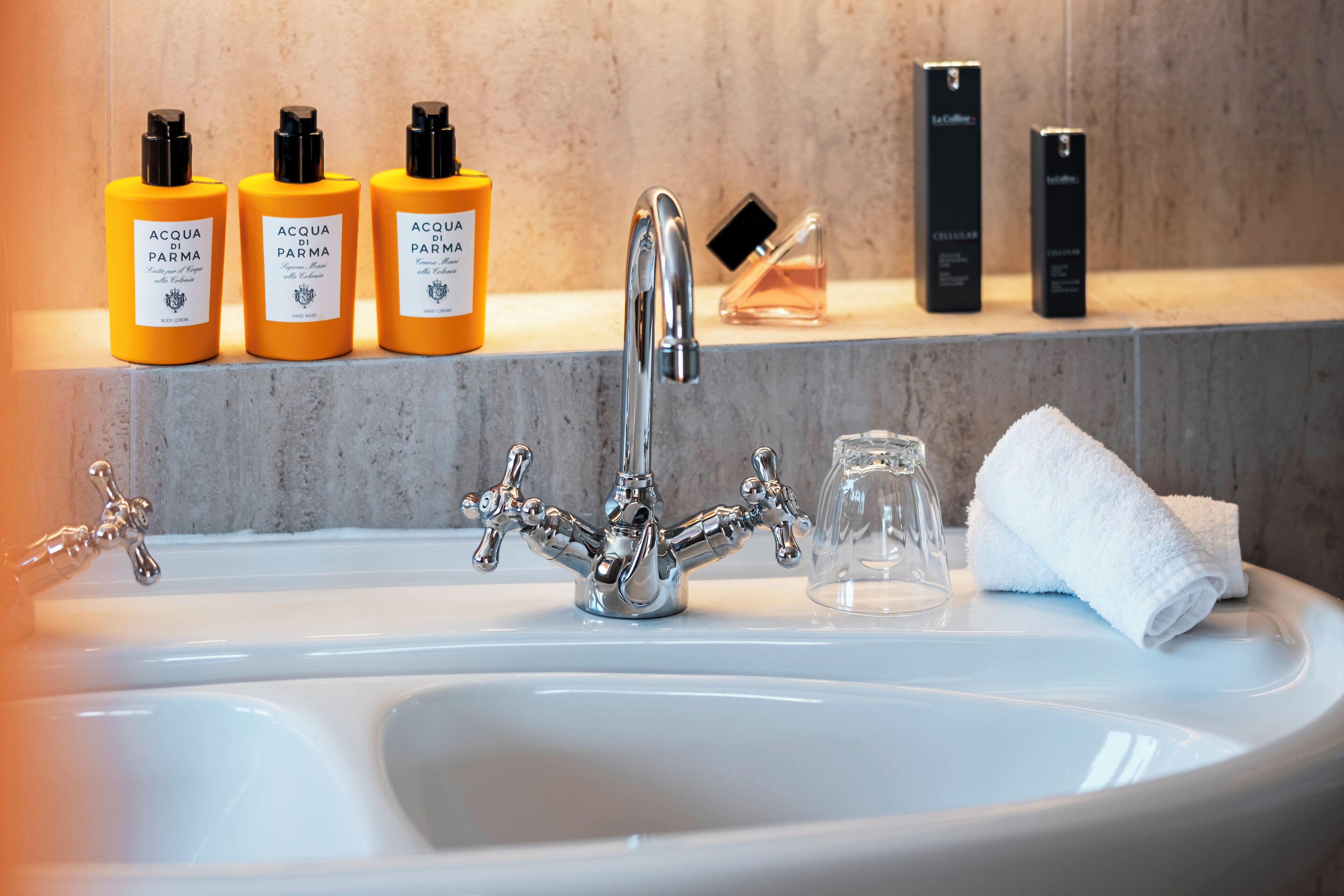 A bathroom sink with a multi-handle faucet, three bottles of Acqua di Parma body lotion, and two perfume bottles on the countertop.