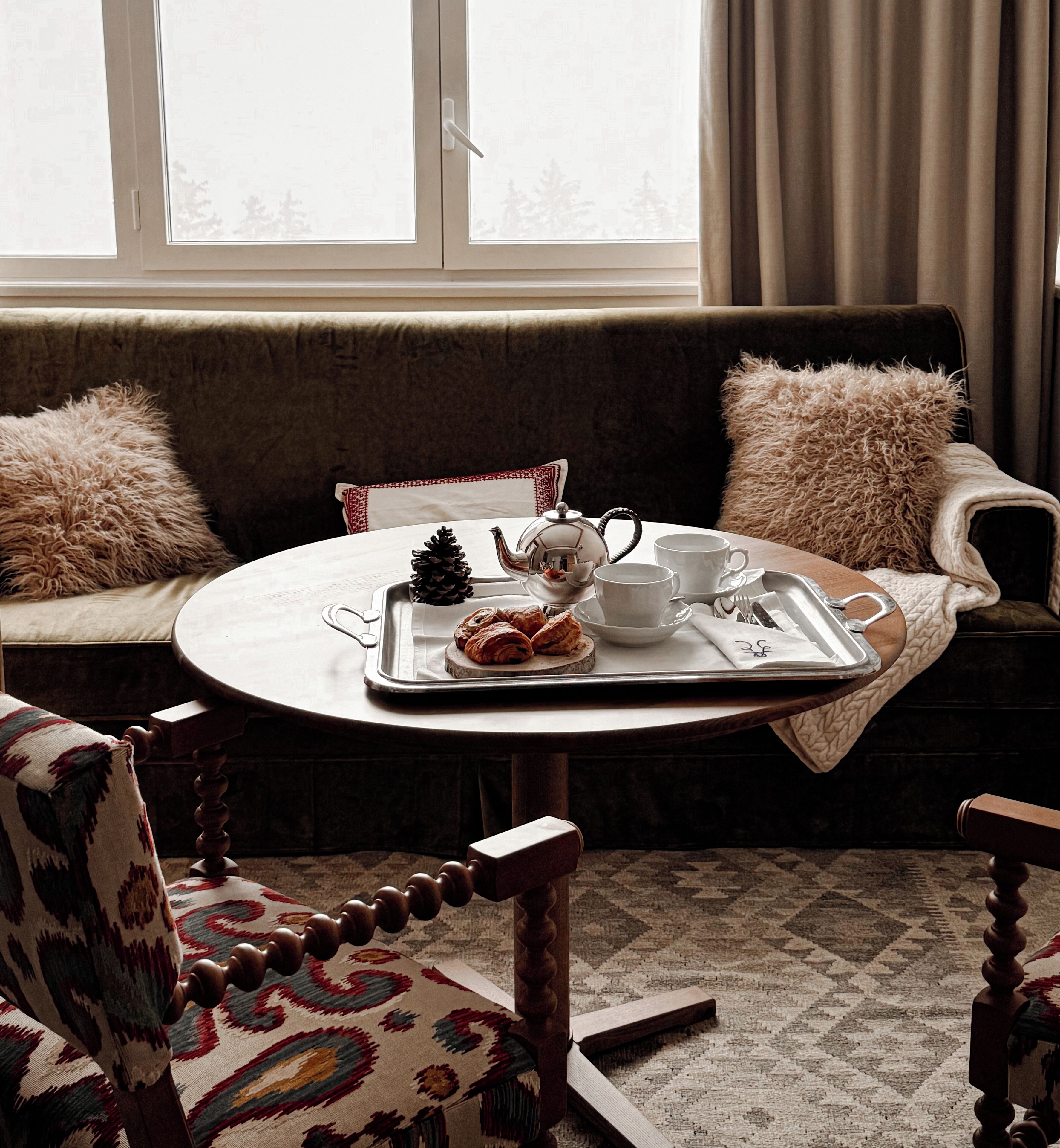 A cozy seating area with a round table set for tea, featuring a tray with pastries, a teapot, cups, and cutlery.
