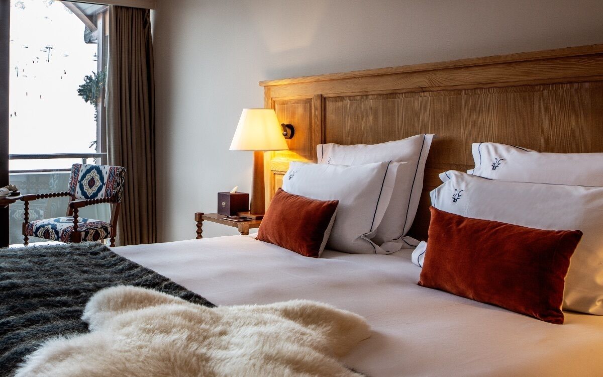 A hotel room with a wooden headboard, a bed with white and brown pillows, a nightstand with a lamp, and a view of the outdoors through a window.