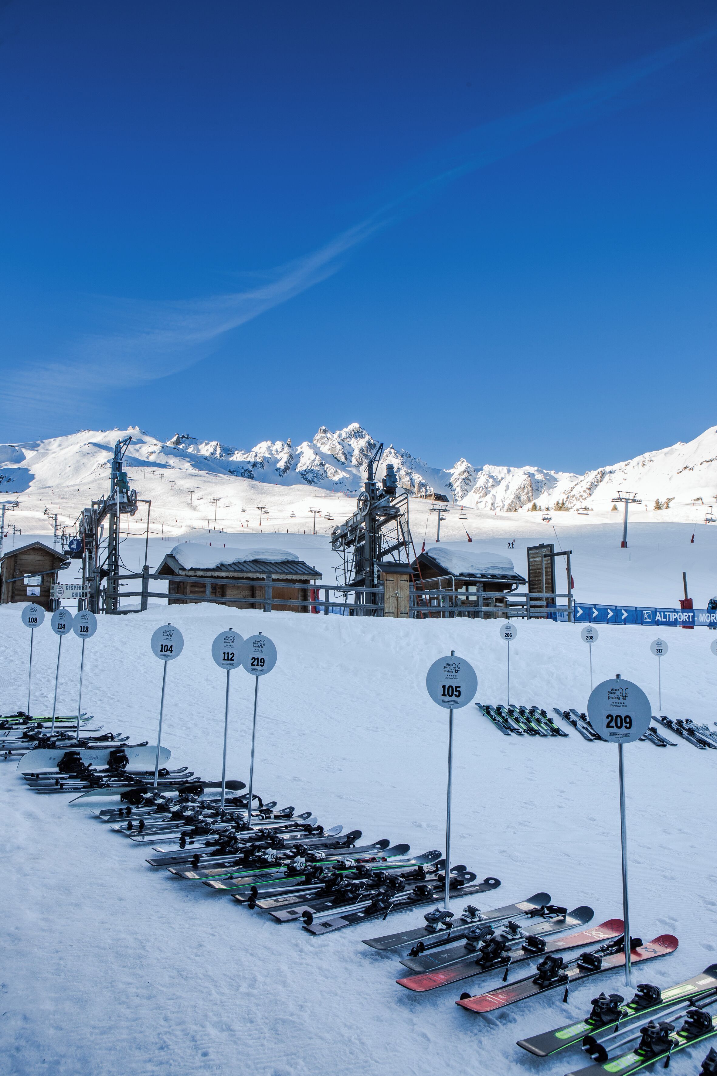 A ski resort with multiple ski lifts, a snow-covered mountain backdrop, and rows of parked skis and snowboards.