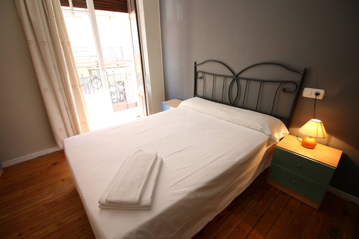 A neatly made bed with a white comforter and a folded towel, a bedside table with a lamp, and a wooden floor.