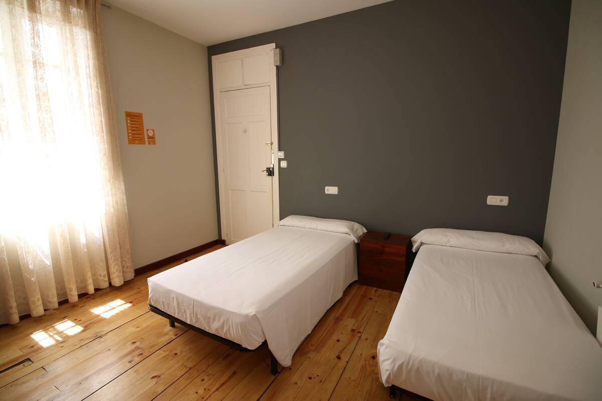 A room with two single beds, wooden flooring, a white door, and a window with curtains.