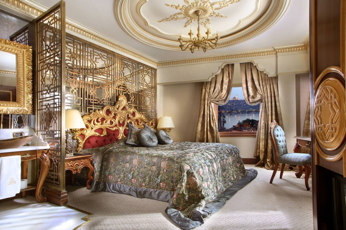 A luxurious bedroom with a large bed, ornate headboard, and a view of a cityscape through a window.