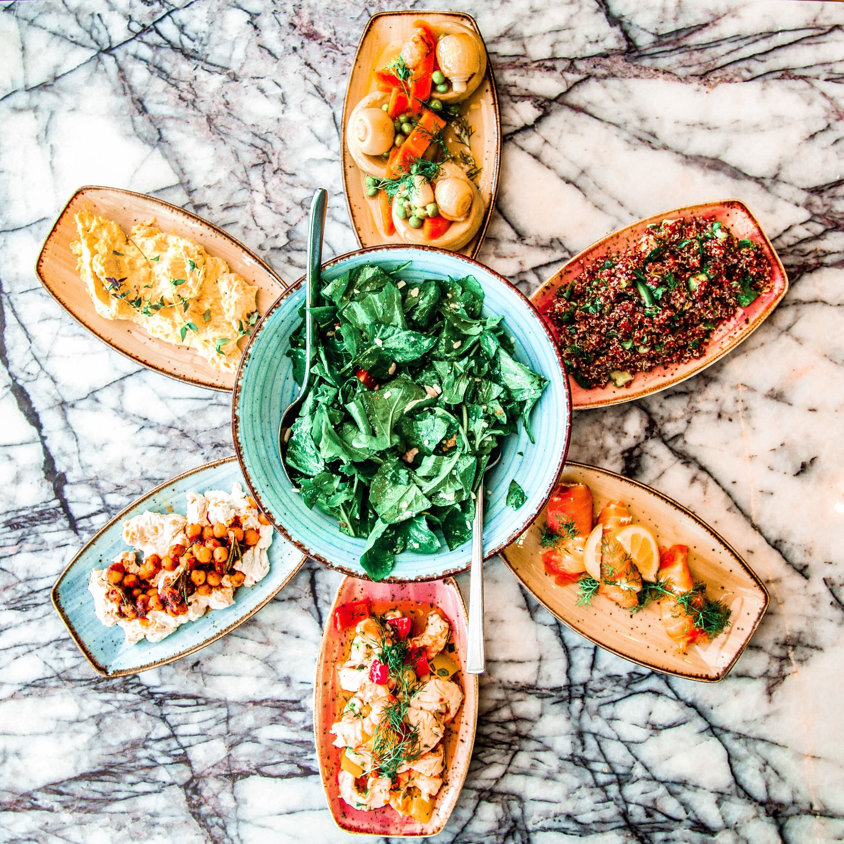 A top-down view of a marble-topped table with six dishes arranged in a circular pattern around a central bowl of greens.