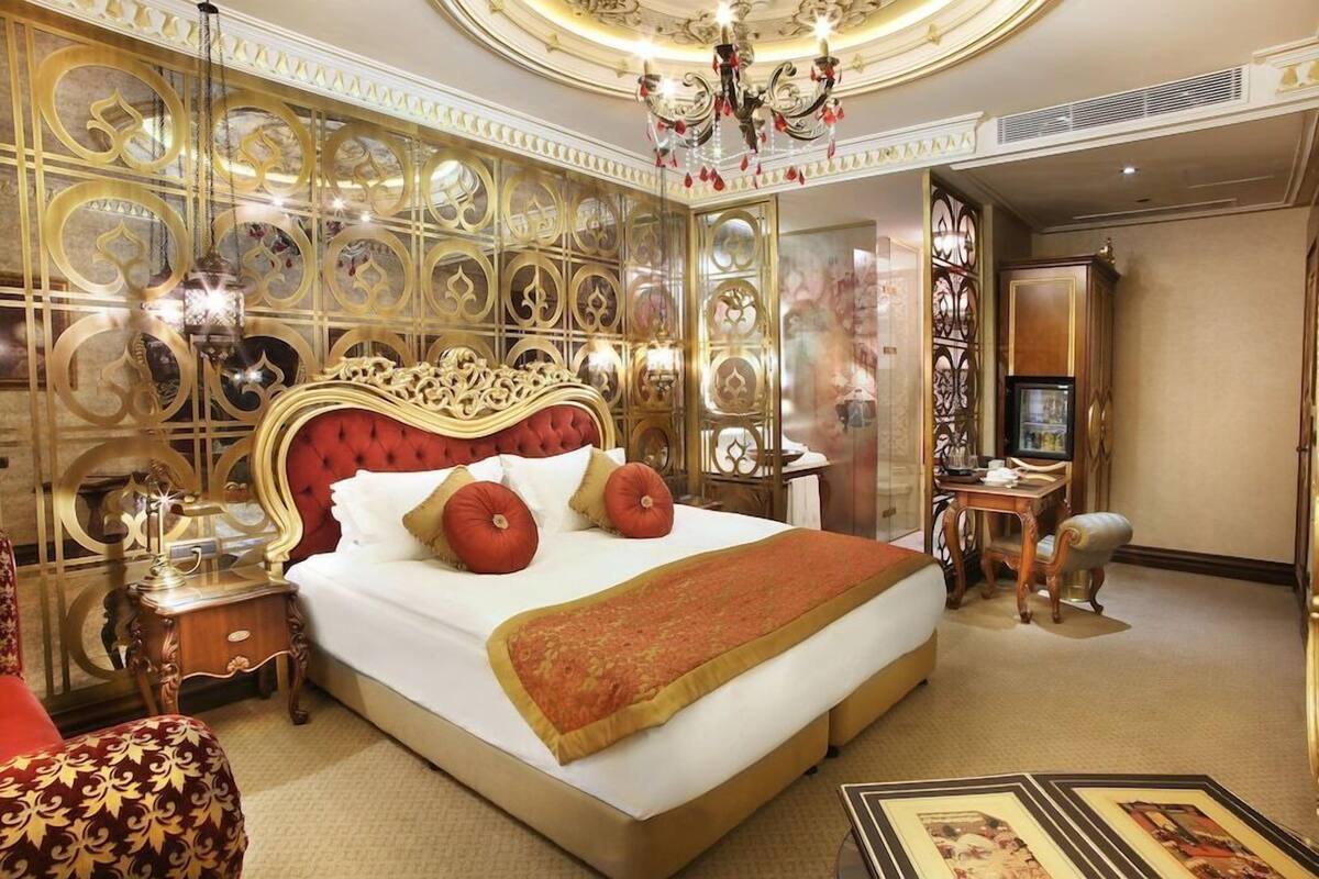 A luxurious hotel room with a large bed, ornate wall paneling, a chandelier, and a desk with a chair.