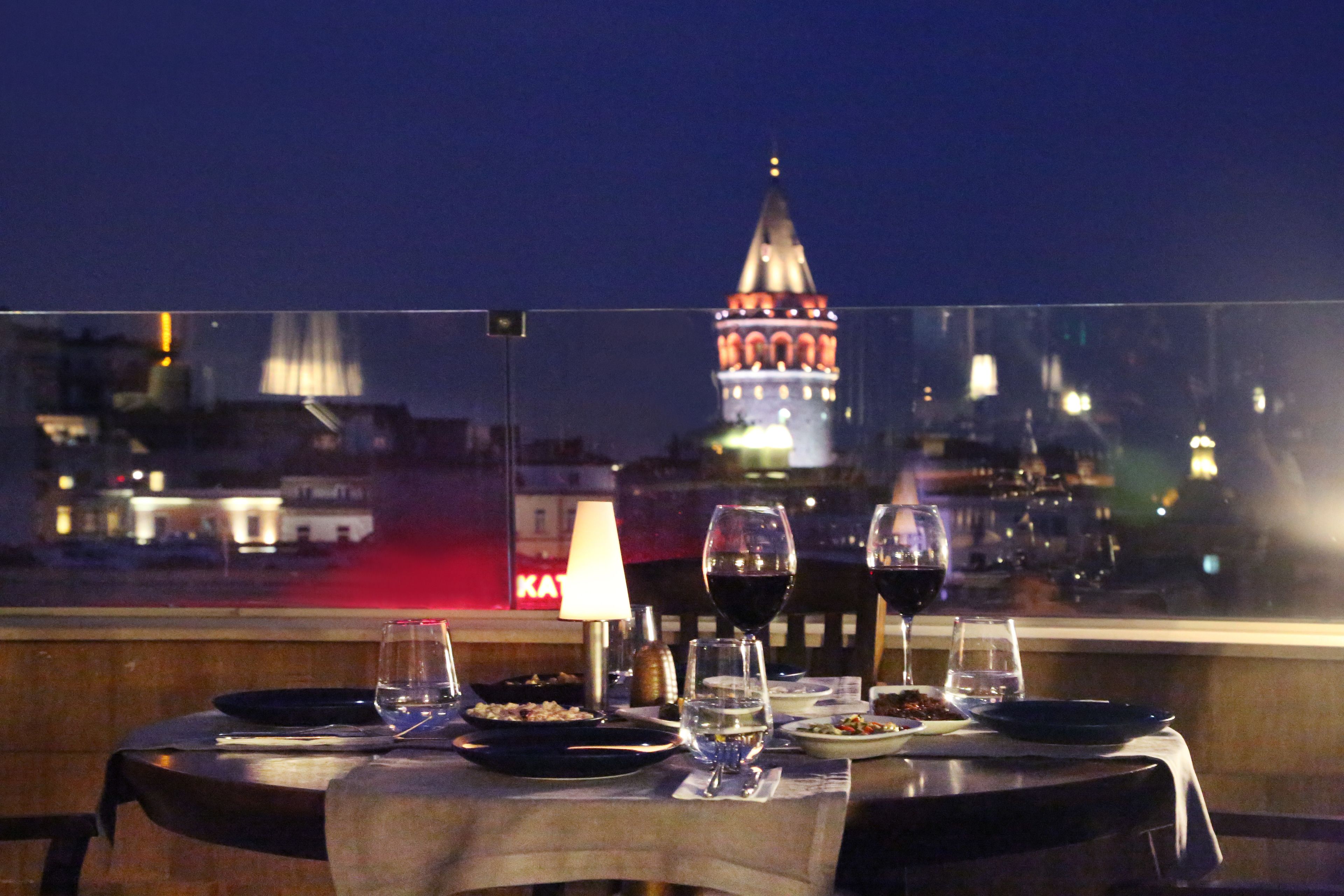 A dining table set with plates, glasses, and a lamp, overlooking a cityscape with a prominent tower.