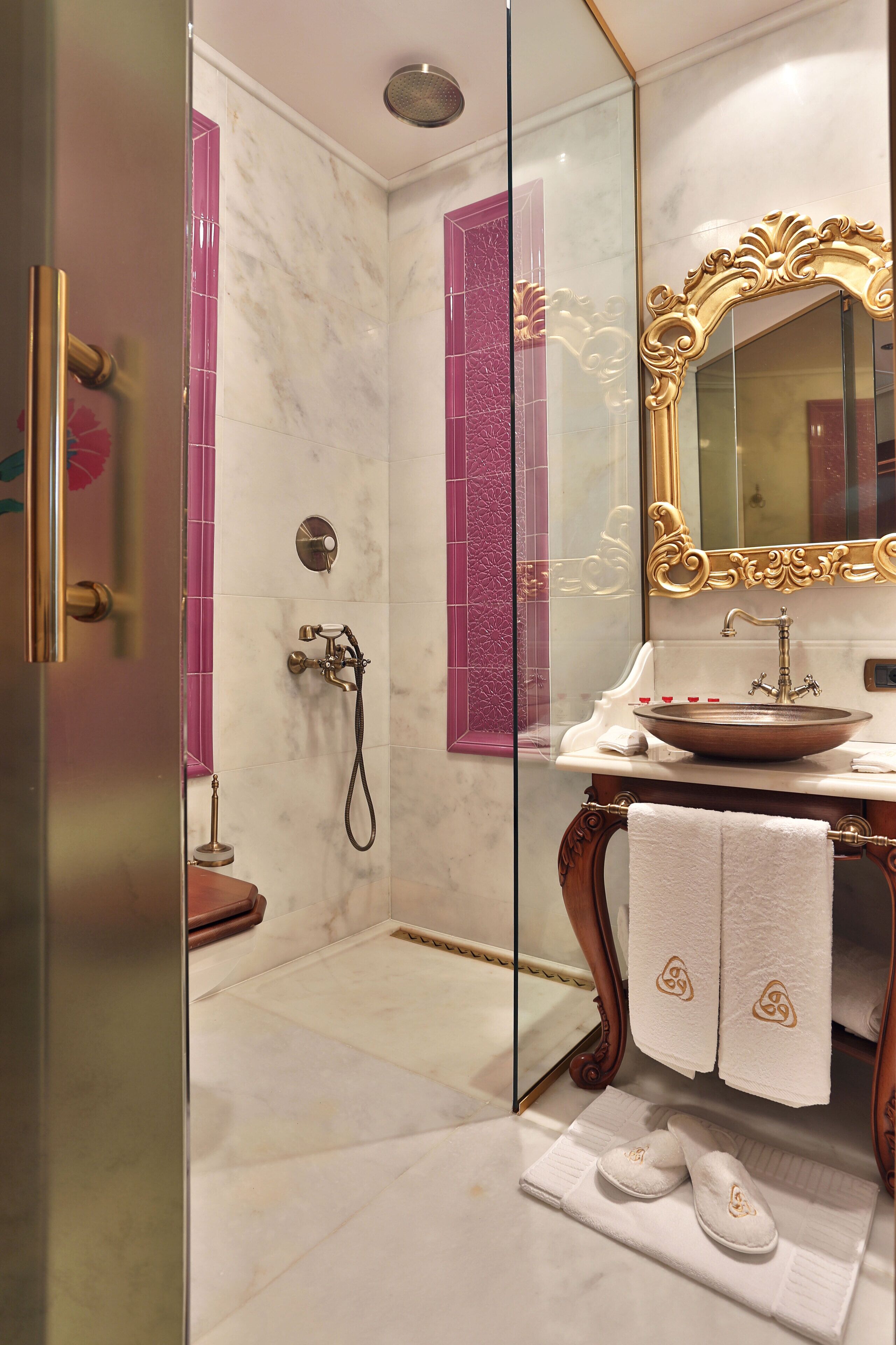 Une salle de bain avec une douche en marbre, un meuble-lavabo doté d’un grand miroir et une paire de chaussons posée sur le sol.