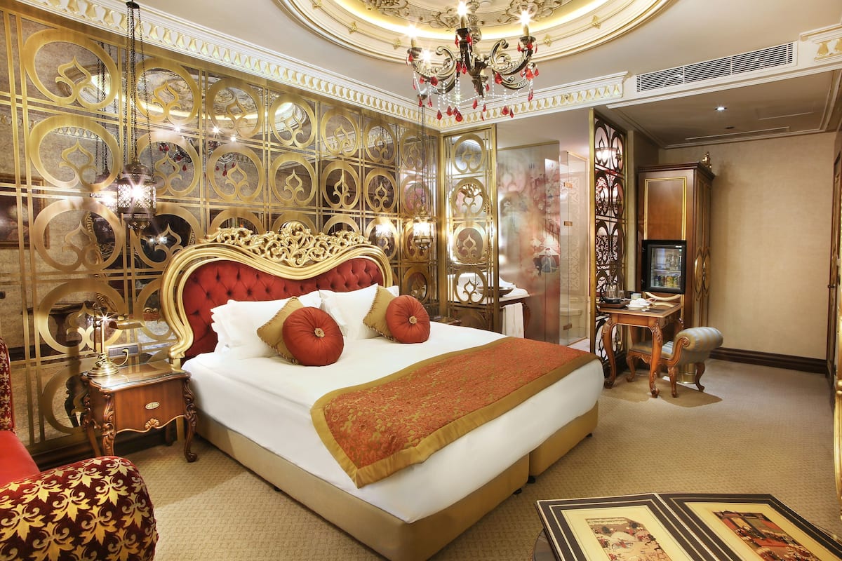 A luxurious hotel room with a large bed, ornate gold wall panels, a chandelier, and a desk with a chair.