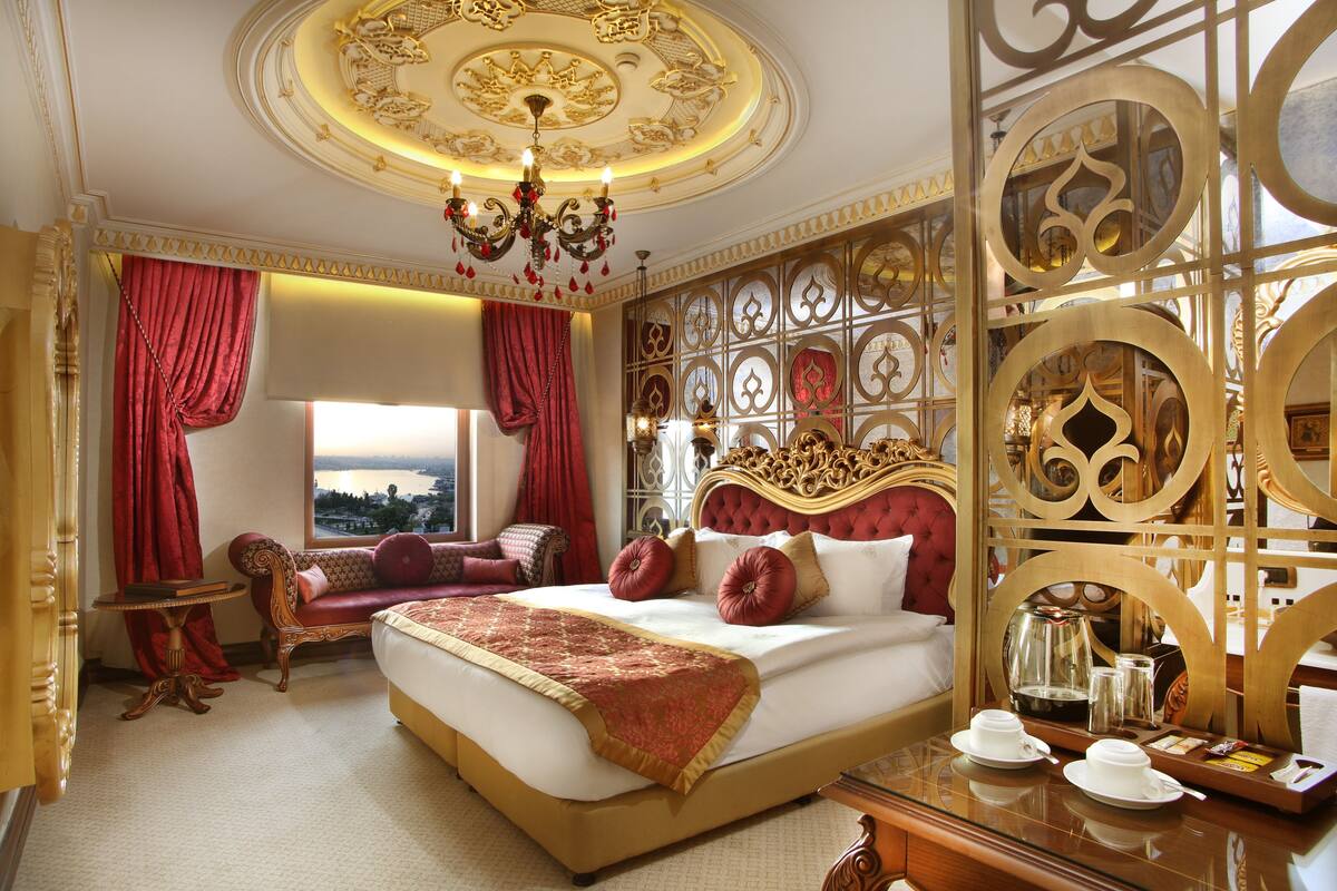 A luxurious hotel room with a large bed, a sitting area with a view, and ornate decor.