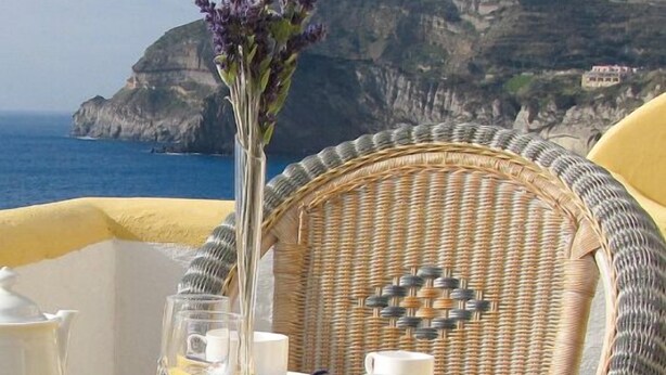 Een gedekte tafel met witte kopjes, schoteltjes en een vaas met paarse bloemen, met uitzicht op een klif aan de kust.