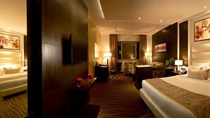 A hotel room with a large bed, a desk, a chair, and a sofa. There are two paintings on the walls and a large window.