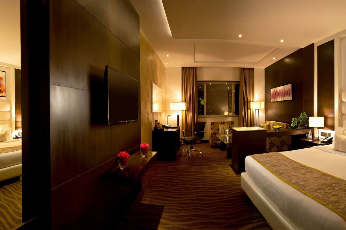 A hotel room with a large bed, a desk, a chair, and a sofa. There are two paintings on the walls and a large window.